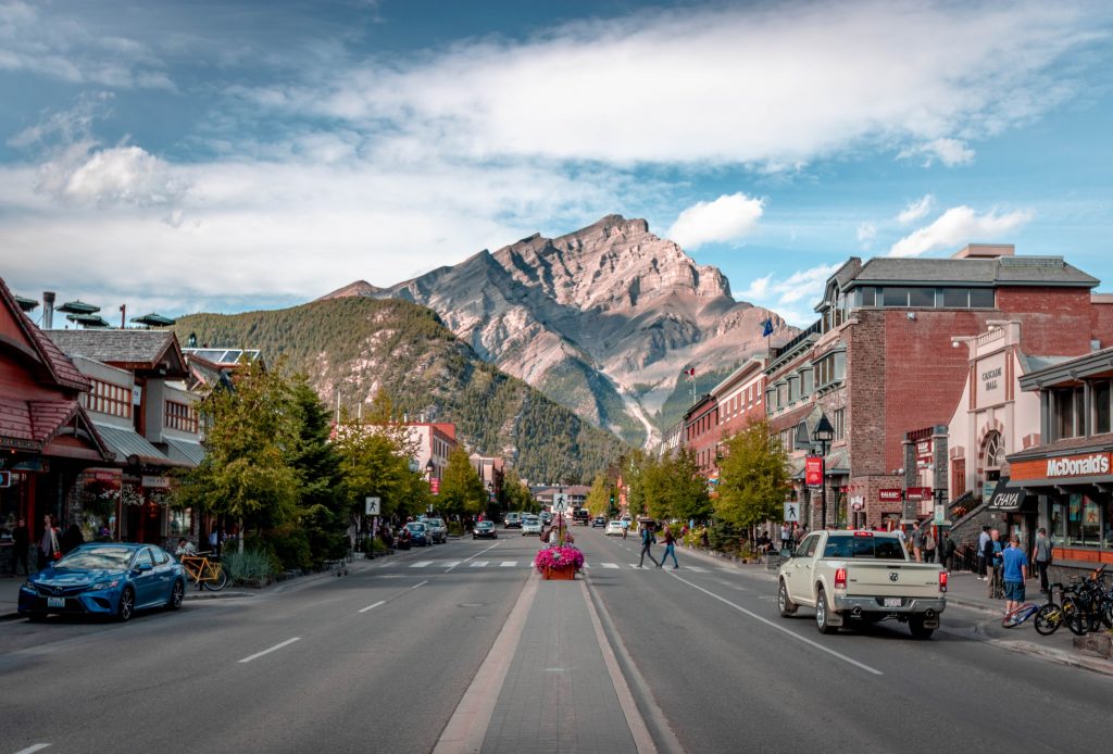 The Rocky Mountains framed by the town of Banff, Alberta. Various vehicles drive towards and away from the camera along the main stretch of road