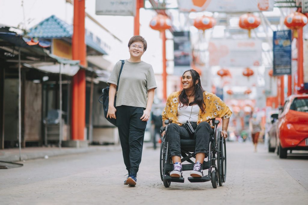 one woman walks beside another in a mobility aid on a city street