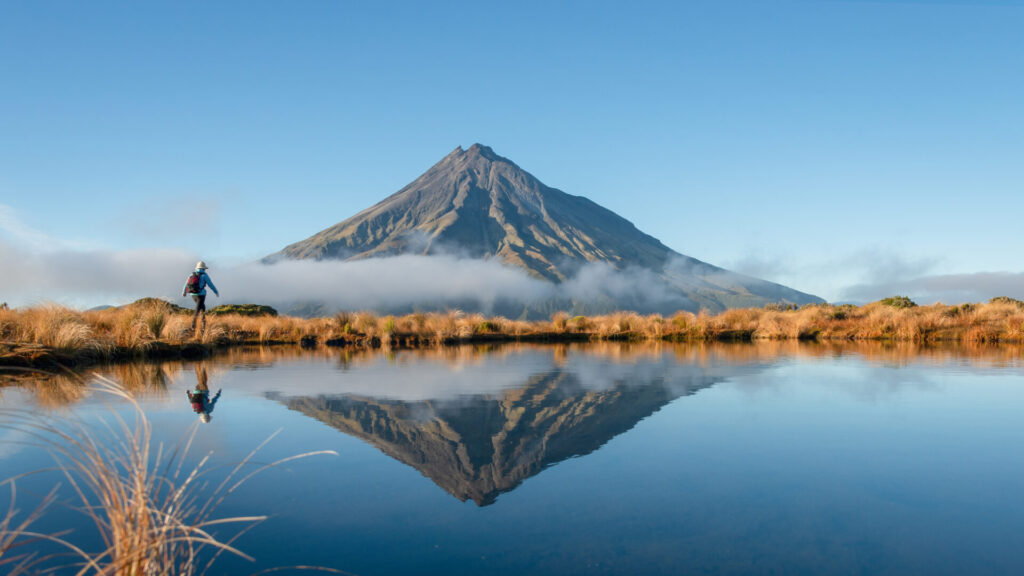 Woman hiking Pouakai circuit. Mt Taranaki reflected in the Pouakai tarn in morning sun. New Zealand.