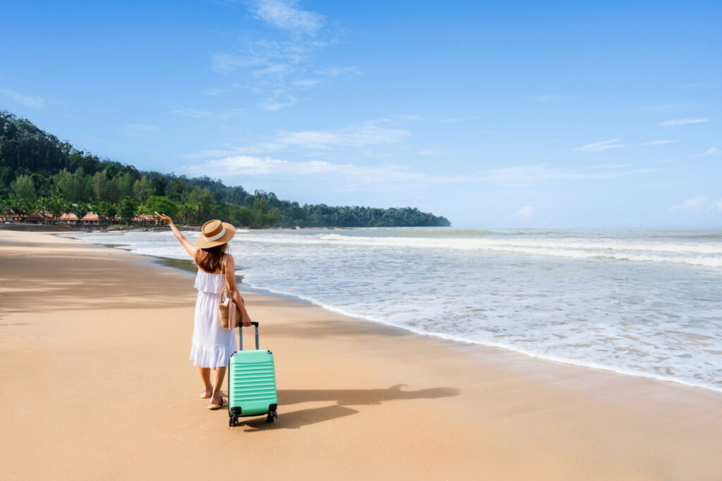 Young woman traveler with luggage relaxing and enjoying on tropical beach in sunny day, Summer vacation and travel concept